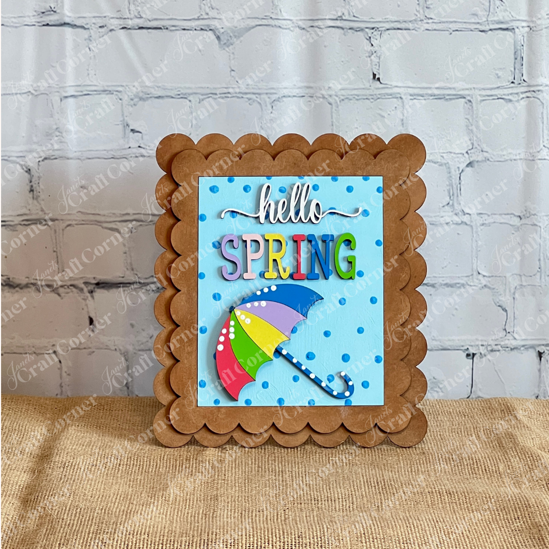 Janet's Craft Corner Spring Switchable Farmhouse Signs DIY kit features "hello SPRING" text and a colorful umbrella on a blue polka dot background with scalloped brown edges—perfect for tiered trays or spring farmhouse decor.