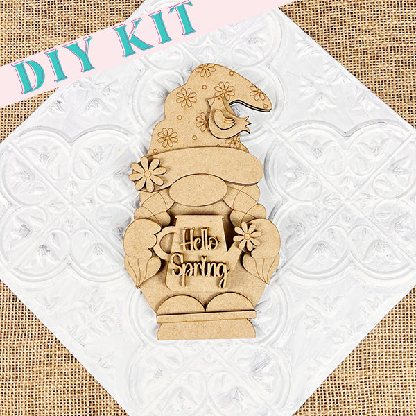 The Hellp Spring Girl Gnome from Janet's Craft Corner features a wooden cutout of a girl gnome wearing a floral hat and holding a mug with the message "Hello Spring." Ideal for spring decor, this charming gnome stands in front of a decorative white textured background, topped with the words "DIY Kit.