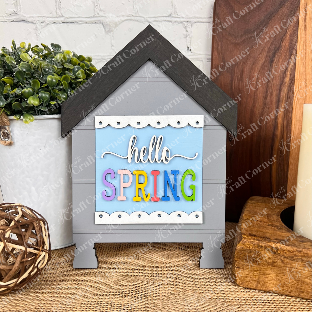 The Hello Spring Squares by Janet's Craft Corner is a decorative gray wooden house with a black roof and colorful "hello SPRING" sign—ideal for tiered tray decor alongside plants, candles, or wicker balls.