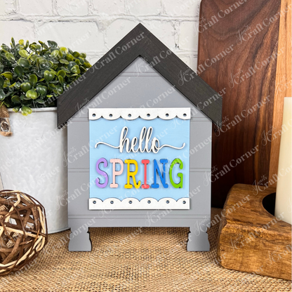 The Hello Spring Squares by Janet's Craft Corner is a decorative gray wooden house with a black roof and colorful "hello SPRING" sign—ideal for tiered tray decor alongside plants, candles, or wicker balls.