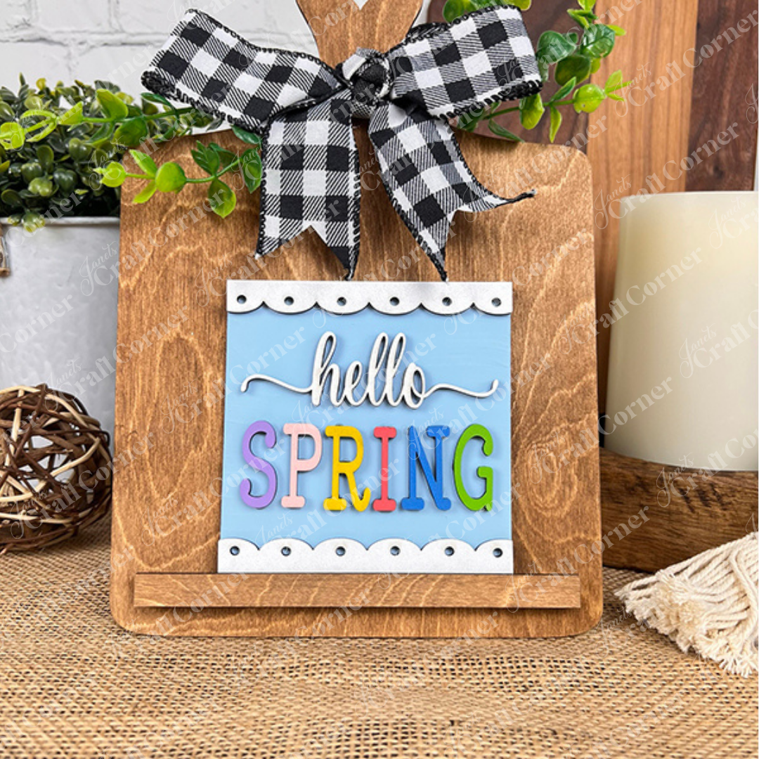 Janet's Craft Corner's "Hello Spring Squares" features a wooden cutting board with a black-and-white checked bow, colorful "hello SPRING" letters, wood cut-outs, and greenery—ideal for tiered tray decor.