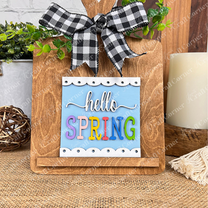 Janet's Craft Corner's "Hello Spring Squares" features a wooden cutting board with a black-and-white checked bow, colorful "hello SPRING" letters, wood cut-outs, and greenery—ideal for tiered tray decor.