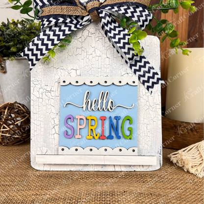 The Hello Spring Squares by Janet's Craft Corner feature playful wood cut-outs, a blue and white background, and vibrant "hello SPRING" letters, topped with a ribbon and greenery—ideal tiered tray decor for any rustic setting.