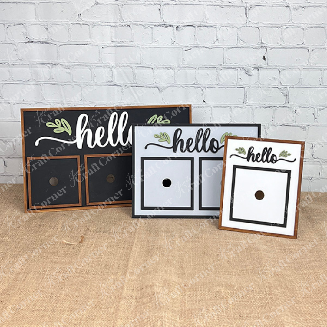Three "Hello Swappable Squares Holder" DIY Craft Kits by Janet's Craft Corner, in black, gray, and white, sit on burlap and feature painted leaves with two spaces for seasonal inserts.