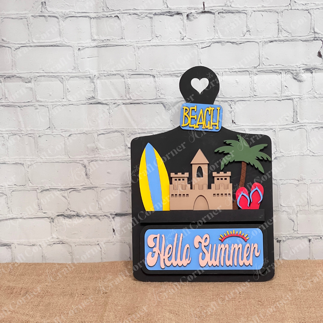 A black breadboard sign by Janet's Craft Corner, from their Summer Beach Interchangeable Insert DIY home decor kit, is set against a brick wall. It features delightful hand-painted images of a sandcastle, surfboard, palm tree, and flip-flops. The scene exudes interchangeable home decor charm with signs reading "Hello Summer" and "Beach." 