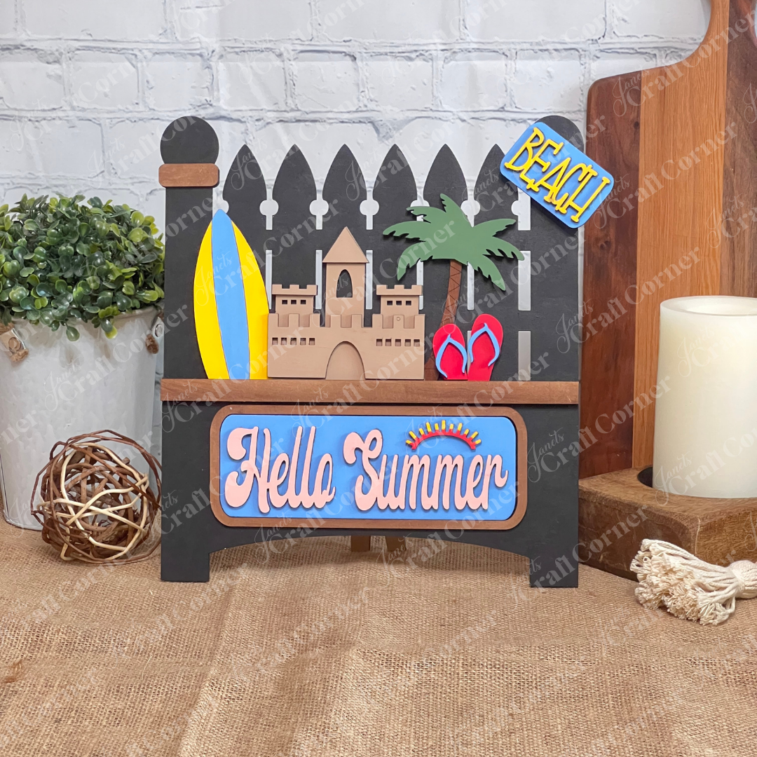 A decorative DIY craft kit sign from Janet's Craft Corner features a surfboard, sandcastle, palm tree, and flip-flops. It reads "Hello Summer" in cursive on a blue background with a yellow "BEACH!" insert against a Picket Fence-inspired brick wall.