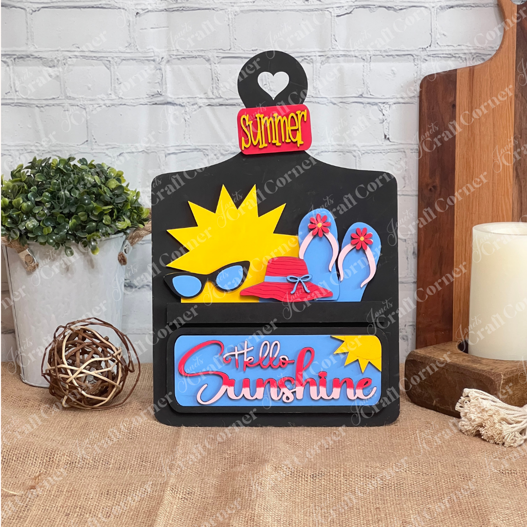 Janet's Craft Corner offers the "Hello Sunshine Interchangeable Insert" craft kit, a wooden card breadboardwith a heart cut-out labeled "Summer." It features postcards with sun and beach motifs, all set against a white brick wall on a brown surface for seasonal home decor.