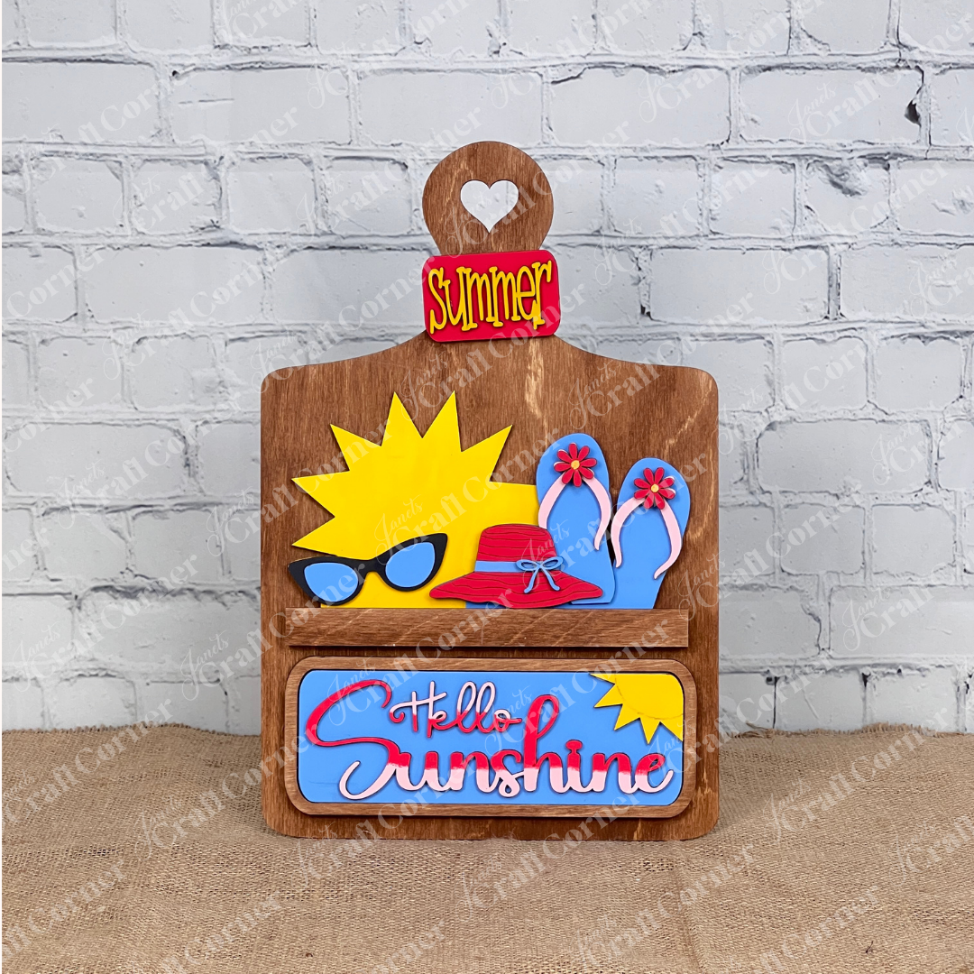 The Hello Sunshine Interchangeable Insert from Janet's Craft Corner includes a wooden decorative sign featuring the word "Summer," a heart cutout, and playful icons like sunglasses and flip-flops, perfect for adding seasonal warmth to your summer picnic against its white brick backdrop.