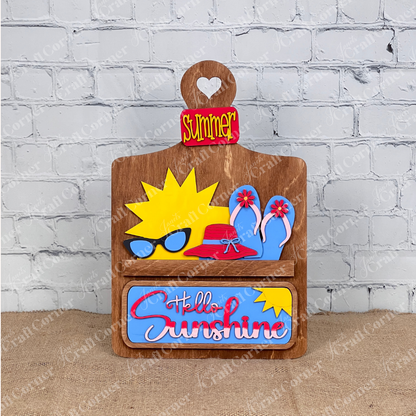 The Hello Sunshine Interchangeable Insert from Janet's Craft Corner includes a wooden decorative sign featuring the word "Summer," a heart cutout, and playful icons like sunglasses and flip-flops, perfect for adding seasonal warmth to your summer picnic against its white brick backdrop.