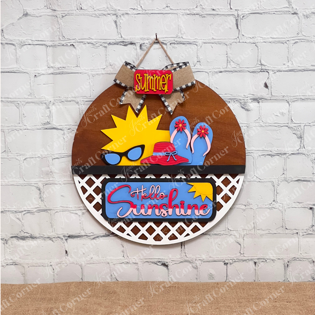 Janet's Craft Corner offers the "Hello Sunshine Interchangeable Insert" craft kit, a round door hanger with lattice work on the bottom third." It features postcards with sun and beach motifs with signs that have the words "summer" and "hello sunshine", all set against a white brick wall on a brown surface for seasonal home decor.