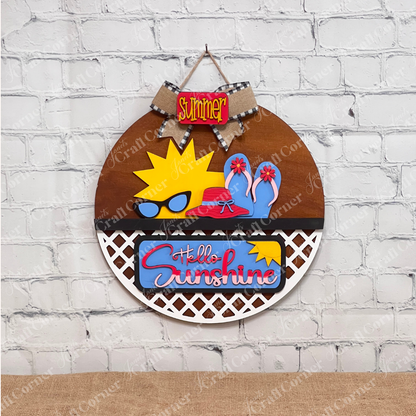 Janet's Craft Corner offers the "Hello Sunshine Interchangeable Insert" craft kit, a round door hanger with lattice work on the bottom third." It features postcards with sun and beach motifs with signs that have the words "summer" and "hello sunshine", all set against a white brick wall on a brown surface for seasonal home decor.