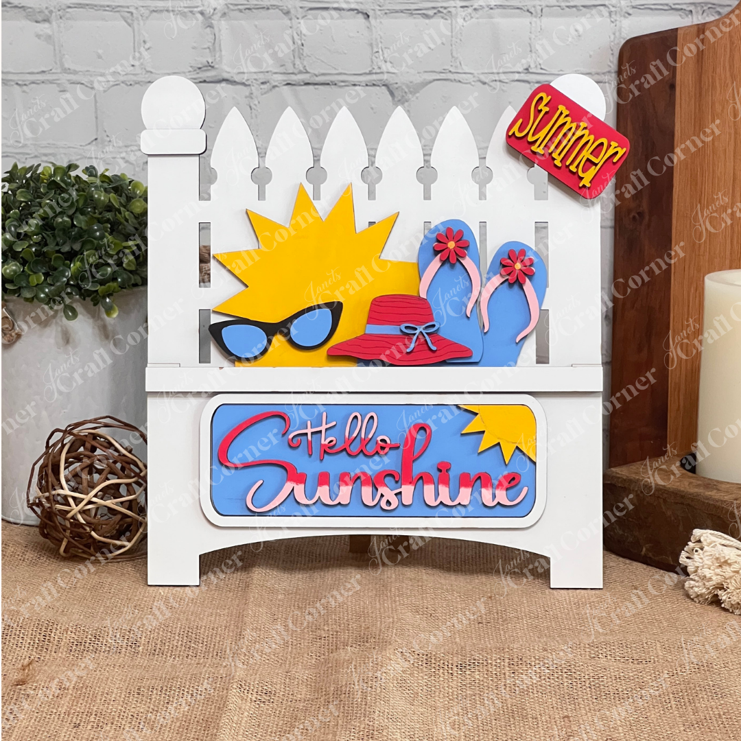 The Hello Sunshine Interchangeable Insert by Janet's Craft Corner is a DIY seasonal decor kit. It includes a white picket fence design featuring a sun with sunglasses and a red hat, flip-flops, "Hello Sunshine" text, and a mini “Summer Picnic” sign against a white brick wall backdrop on burlap.