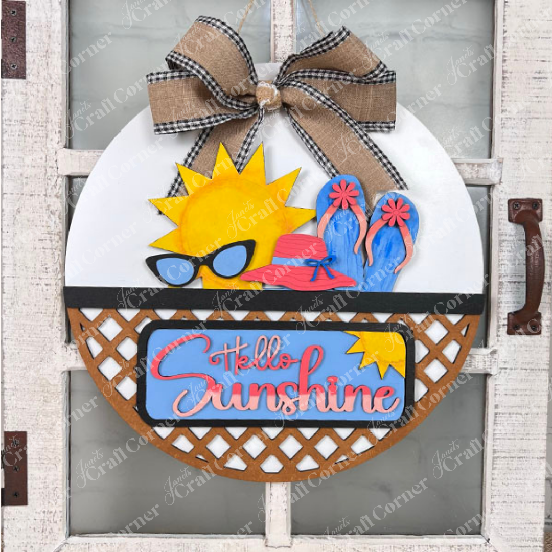 Janet's Craft Corner offers the "Hello Sunshine Interchangeable Insert" craft kit, a round door hanger with stained lattice work on the bottom third." It features postcards with sun and beach motifs with signs that have the words "summer" and "hello sunshine", all set against a white brick wall on a brown surface for seasonal home decor.
