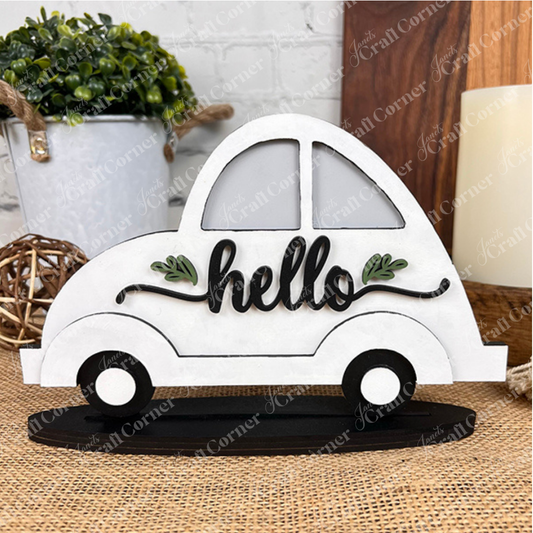 A white wooden “Hello Volkswagen - Painted” car shelf sitter by Heartland, featuring the word “hello” and green leaves, is displayed on a black base. Bringing a touch of country charm, a plant, wicker ball, and candle sit in the background on a textured surface.