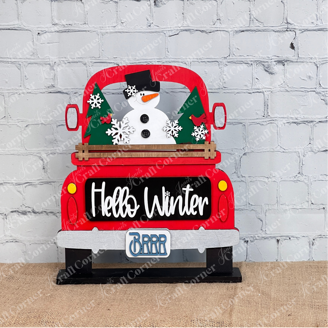 The Hello Winter Interchangeable Insert by Janet's Craft Corner is a DIY seasonal home decor craft kit featuring a red pickup with a snowman, trees, and snowflakes. The tailgate says "Hello Winter" and the license plate reads "BRRR." Set against a white brick wall, it includes 3 charming pieces.