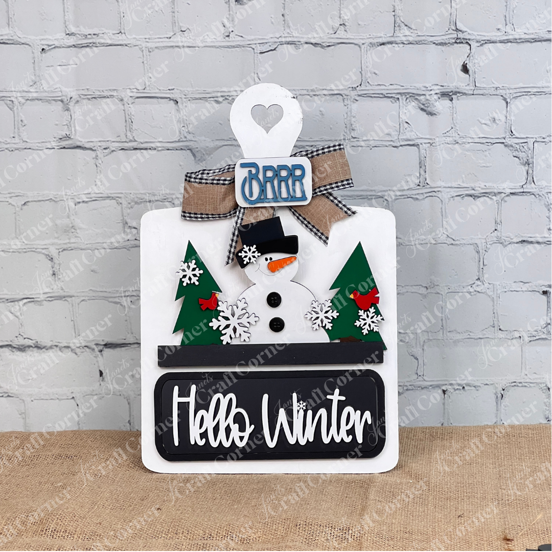 The Hello Winter Interchangeable Insert by Janet's Craft Corner features a cheerful snowman with a black hat and red scarf, flanked by green trees. Snowflakes, red birds, and a burlap bow adorn the "Brrr" sign, with "Hello Winter" on a plaque. Ideal for white brick walls as DIY seasonal decor.