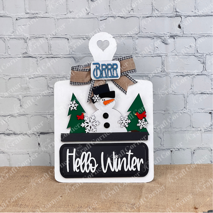 The Hello Winter Interchangeable Insert by Janet's Craft Corner features a cheerful snowman with a black hat and red scarf, flanked by green trees. Snowflakes, red birds, and a burlap bow adorn the "Brrr" sign, with "Hello Winter" on a plaque. Ideal for white brick walls as DIY seasonal decor.