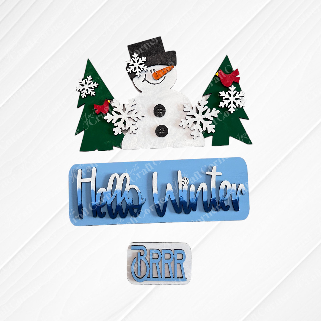 Janet's Craft Corner's Hello Winter Interchangeable Insert is a DIY seasonal home decor kit featuring a smiling snowman in a top hat with green trees, red birds, and blue "Hello Winter" and “Brrr” text on textured white—set includes 3 pieces.