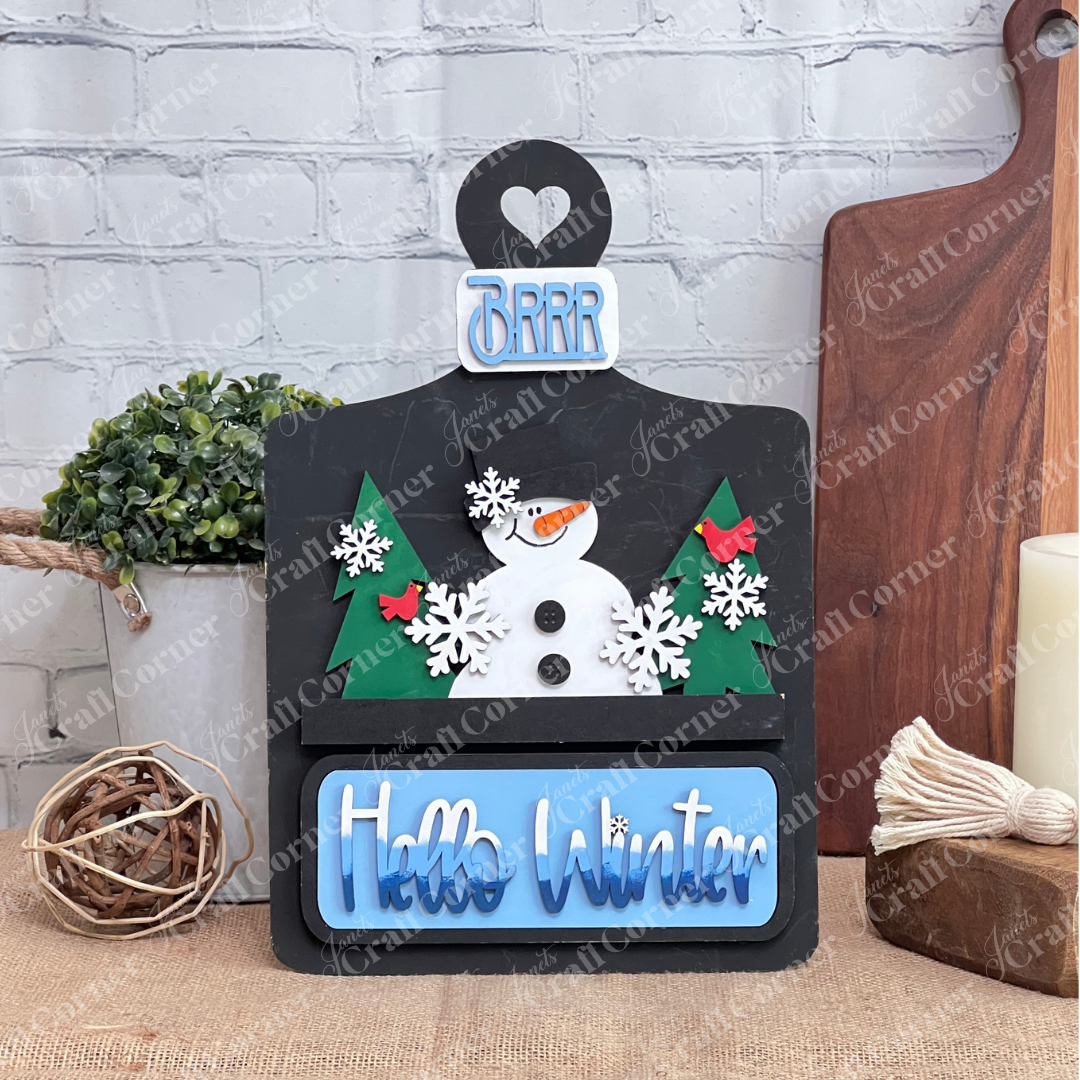The Hello Winter Interchangeable Insert by Janet's Craft Corner is a DIY seasonal home decor craft kit featuring a black breadboard with a snowman, trees, and snowflakes. The tailgate has white/blue gradient "Hello Winter" and the license plate has light blue “Brrr” text Set against a white brick wall, it includes 3 charming pieces.