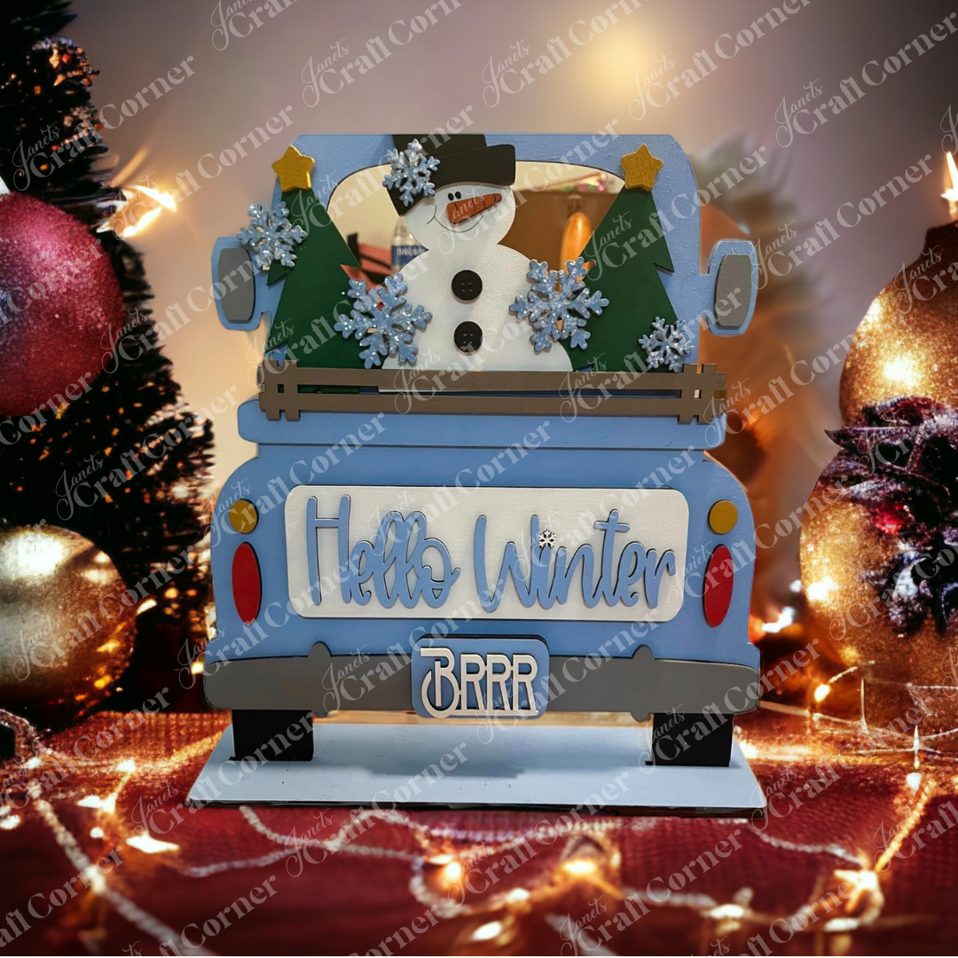 Create a festive holiday scene with Janet's Craft Corner's "Hello Winter" Interchangeable Insert - Painted. This charming decor piece features an antique blue truck adorned with "Hello Winter" on the back, surrounded by a delightful snowman, trees, snowflakes, and Christmas elements such as ornaments and string lights. Designed to offer interchangeable inserts for seasonal flair.