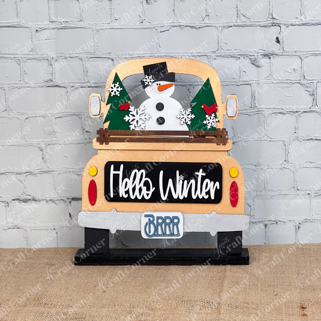 The "Hello Winter" interchangeable insert from Janet's Craft Corner is a DIY seasonal decor kit featuring a truck with swappable inserts of a snowman, trees, and snowflakes. The truck displays "Hello Winter" and "BRRR" on its license plate, set against a burlap surface by a white brick wall for cozy vibes.