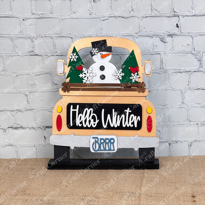 The "Hello Winter" interchangeable insert from Janet's Craft Corner is a DIY seasonal decor kit featuring a truck with swappable inserts of a snowman, trees, and snowflakes. The truck displays "Hello Winter" and "BRRR" on its license plate, set against a burlap surface by a white brick wall for cozy vibes.