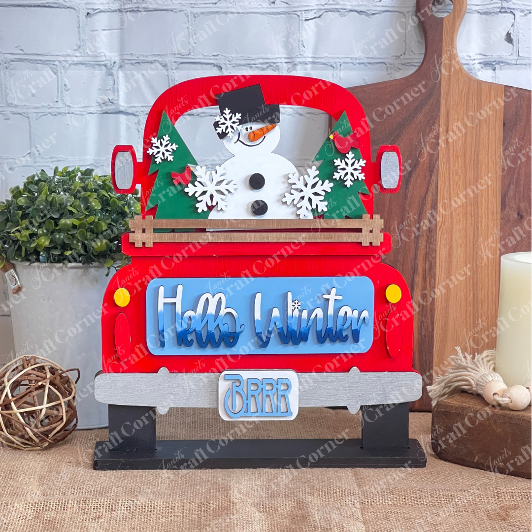 The Hello Winter Interchangeable Insert by Janet's Craft Corner is a DIY seasonal home decor craft kit featuring a red pickup with a snowman, trees, and snowflakes. The tailgate has white/blue gradiant "Hello Winter" and the license plate has light blue “Brrr” text Set against a white brick wall, it includes 3 charming pieces.