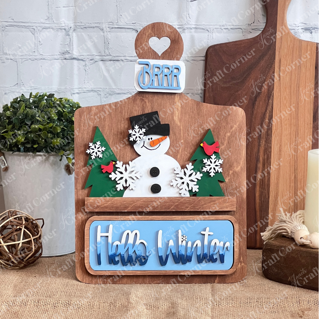 Janet's Craft Corner offers a DIY Breadboard Interchangeable Base with Insert, featuring a snowman, green trees, snowflakes, red birds, a "Brrr" tag bow, and "Hello Winter" text. It's perfect for creative decor enthusiasts to craft cozy home decor against a white brick wall.