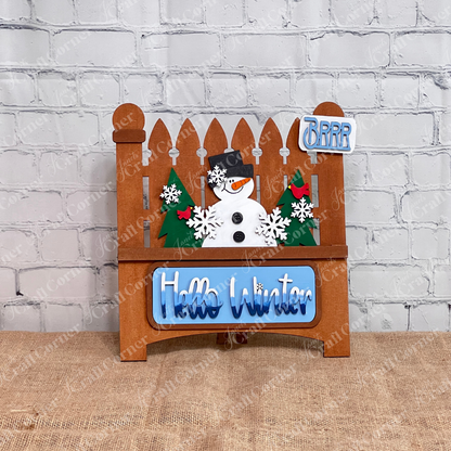 The Hello Winter Interchangeable Insert by Janet's Craft Corner is a DIY seasonal home decor craft kit featuring a stained garden fence with a snowman, trees, and snowflakes. The tailgate has white/blue gradient "Hello Winter" and the license plate has light blue “Brrr” text Set against a white brick wall, it includes 3 charming pieces.