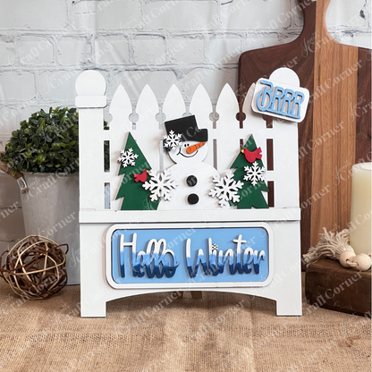 The Hello Winter Interchangeable Insert by Janet's Craft Corner is a DIY seasonal home decor craft kit featuring a white garden fence with a snowman, trees, and snowflakes. The tailgate has white/blue gradient "Hello Winter" and the license plate has light blue “Brrr” text Set against a white brick wall, it includes 3 charming pieces.