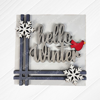 The "Hello Winter Squares" DIY craft kit from Janet's Craft Corner features "hello Winter" in cursive with two white snowflakes and a red cardinal on a gray and blue background—perfect for charming seasonal home decor.