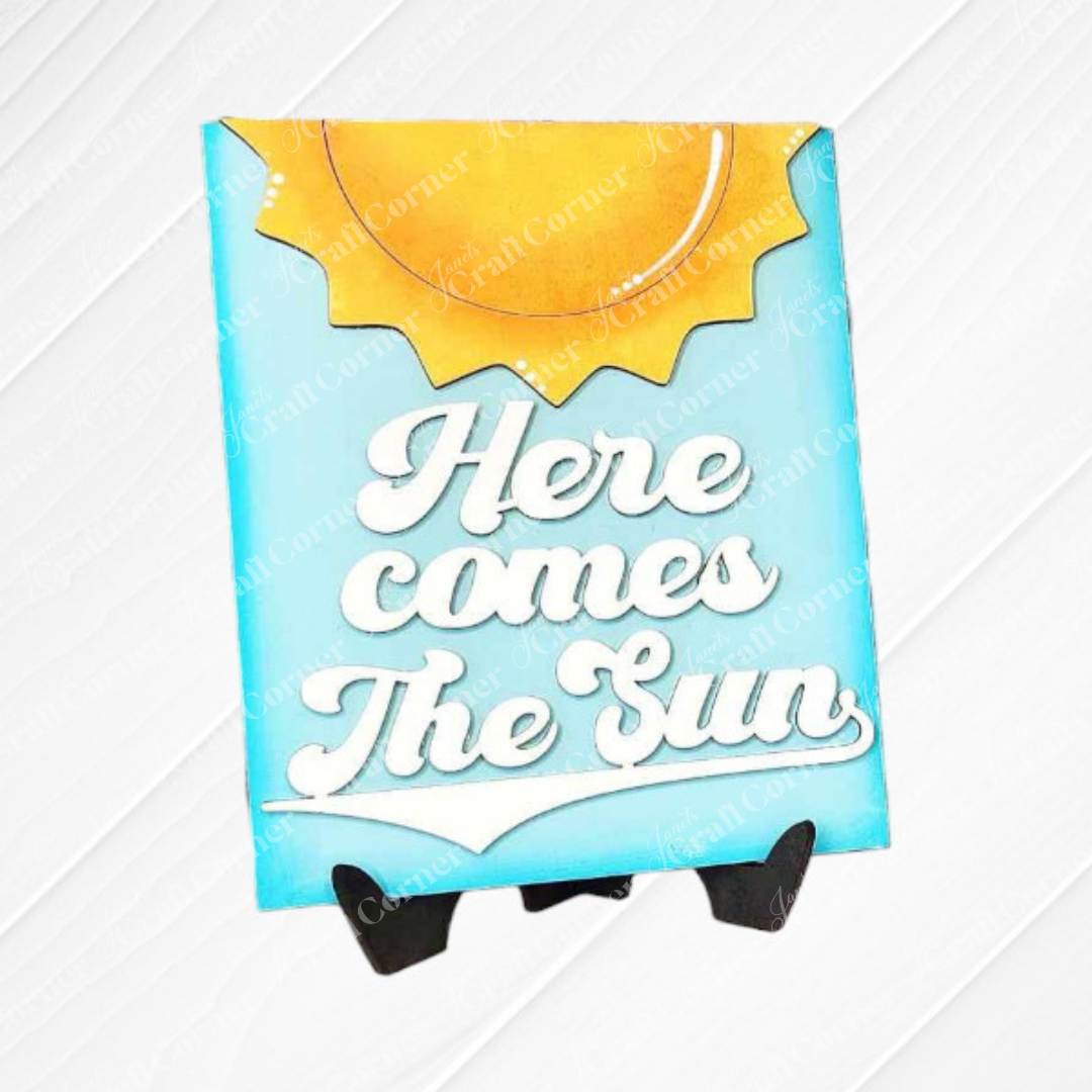 Janet's Craft Corner Summer Switchable Farmhouse Sign features “Here comes The Sun” in white cursive on a yellow sun and blue sky background. This DIY decor kit is perfect for rustic style, displayed on a small black stand. Set of 1.