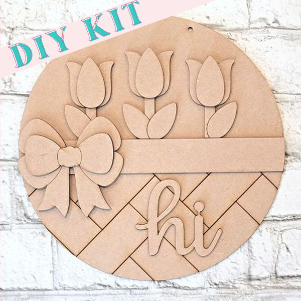 The "Hi Tulip Round" by Janet's Craft Corner provides a delightful enhancement to your home décor. This circular DIY kit includes three tulip shapes, a bow, and the word "hi" set against a woven-patterned background. Ideal for craft enthusiasts, it's beautifully displayed against a white brick wall.