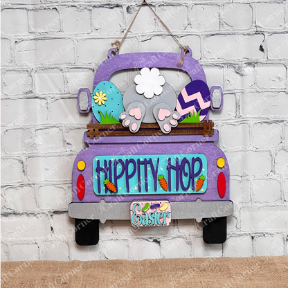 The Antique Truck Base with Insert DIY kit by Janet's Craft Corner features a wooden truck with bunny ears and tail, adorned with colorful eggs and flowers. It displays "HIPPITY HOP" and "Easter," set on burlap against a white brick wall, offering charming hand-painted decor.