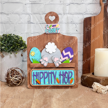 Craft an Easter-themed decoration with Janet's Craft Corner DIY kit. The kit includes a Breadboard Interchangeable Base and Insert featuring a bunny's rear and decorated eggs. "Hippity Hop" text with small carrots pops against a brick background, perfect for fun home decor for all ages!.