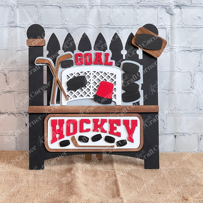 The Hockey Interchangeable Insert from Janet's Craft Corner is a DIY seasonal home decor craft kit featuring a sleek, black background and includes 3 pieces: a goal net, crossed sticks, and multiple black pucks with "Goal" and "Hockey" in striking red and white text.