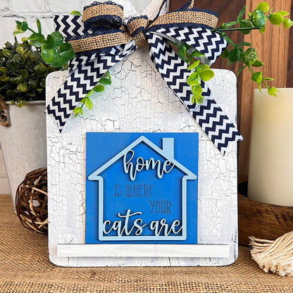 The Cat Swappable Squares by Janet's Craft Corner is a blue house-shaped decorative sign with "Home is where your cats are," a black-and-white chevron ribbon, and greenery on a rustic white background, perfect for tiered trays.