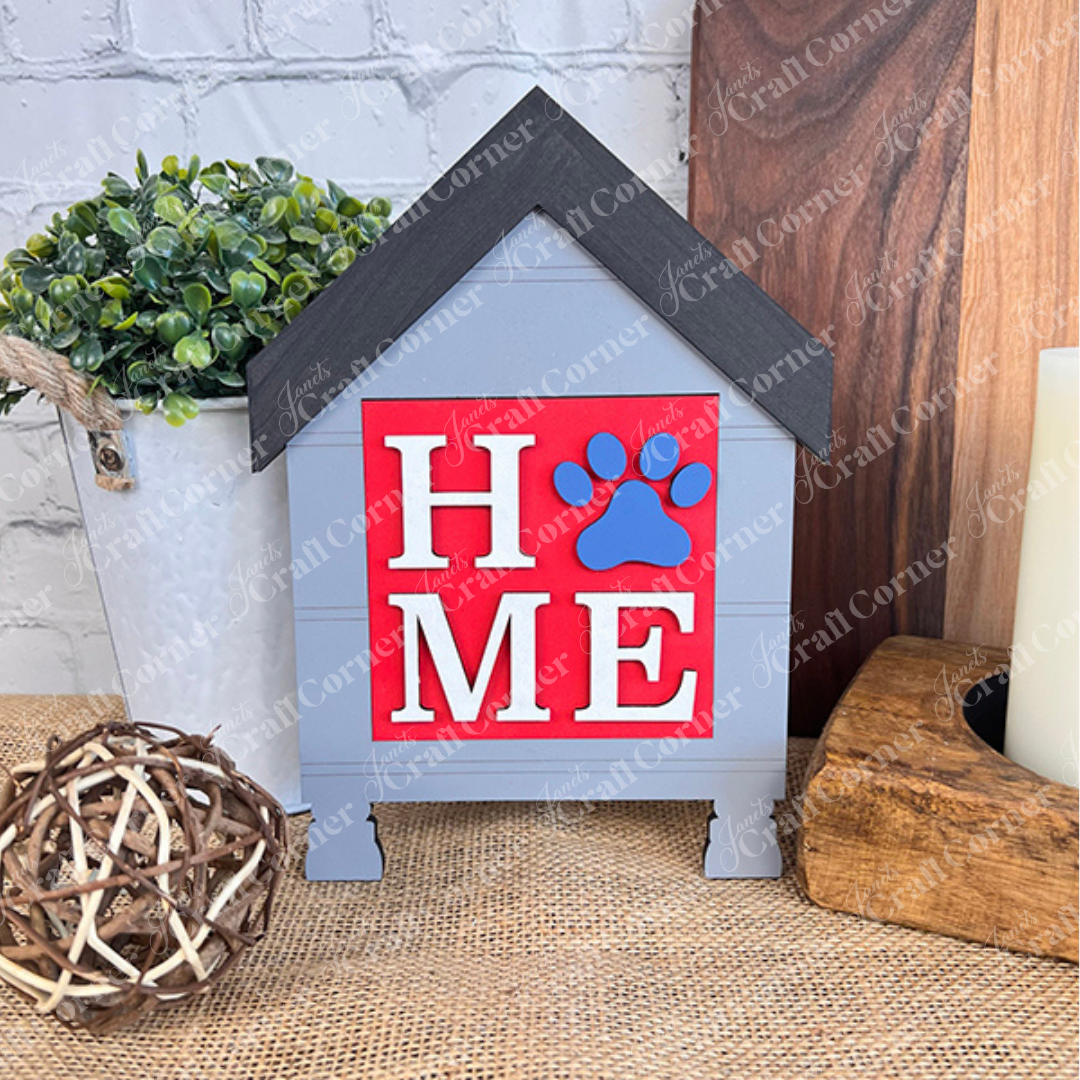 The Dog Squares by Janet's Craft Corner feature a decorative wood sign shaped like a doghouse, gray with a black roof. "HOME" is displayed with a blue paw print as the "O," making it perfect tiered tray decor for pet lovers.