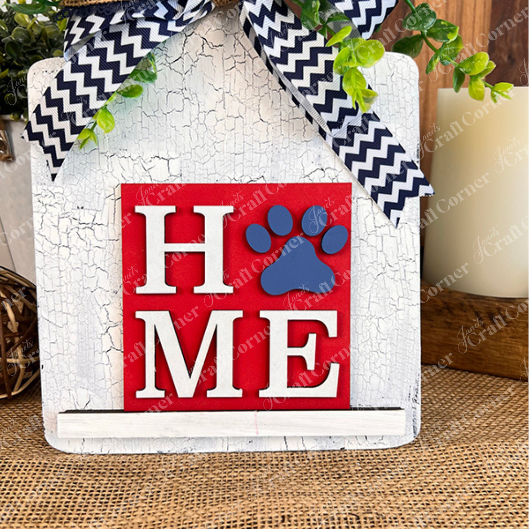 Dog Squares by Janet's Craft Corner features "HOME" on a rustic white wooden board, with the "O" as a blue paw print on red. The design includes wood cut-outs, black-and-white chevron ribbon, and greenery accents.