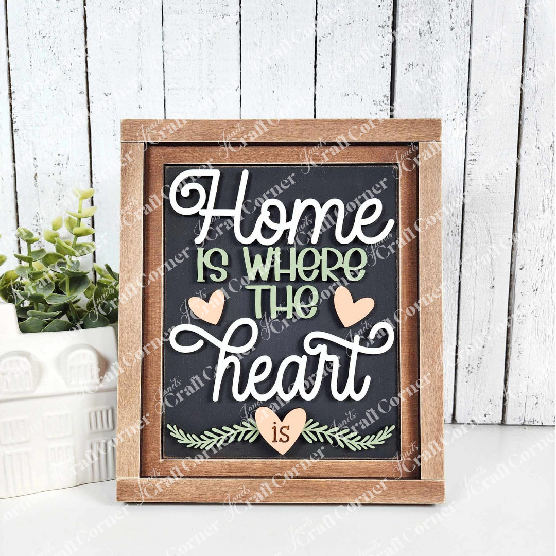 Janet's Craft Corner's Home Heart Interchangeable Sign reads "Home is where the heart is". In this sample, it is done in stylized white and green text with small hearts. Perfect for home décor, it’s propped beside a plant and ceramic house on a light wooden background, adding charm to any space.