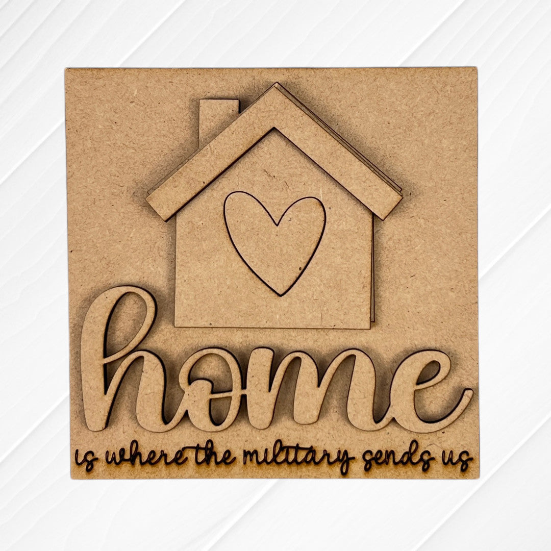 The Military Squares sign by Janet's Craft Corner features a house with a heart cutout and “home” in script, plus “is where the military sends us,” perfect for tiered tray decor with wooden accents on a light background.