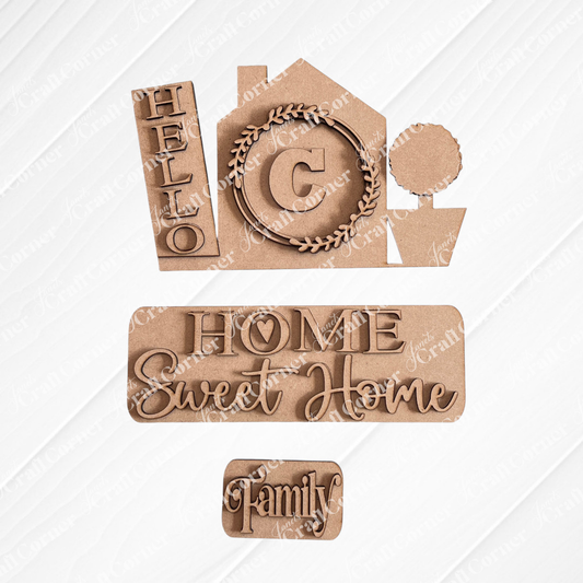 Crafted by Janet's Craft Corner, the Hello Home Monogram Interchangeable Insert | DIY home decor kit features wooden cutouts on a textured surface displaying a house with the word "Hello" and an interchangeable monogram insert, including a "C" wreath. Accompanied by accents like a potted tree and words such as "Home Sweet Home" and "Family," this scene provides a hand-painted DIY feel against the patterned fabric backdrop.