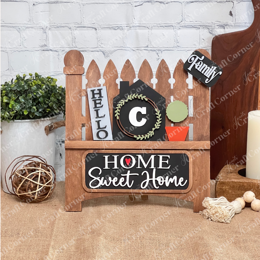 A stained garden fence on a burlap covered shelf in front of a brick wall features a small wreath, "C," and phrases like "Family" in various fonts. This charming piece doubles as Janet's Craft Corner Home Monogram Interchangeable Insert DIY home decor craft kit, featuring 3 pieces.