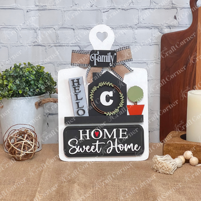 The Breadboard Interchangeable Base with Insert by Janet's Craft Corner features a heart-topped sign with "Family," "Hello," a wreath, a potted plant, and "Home Sweet Home" in bold on burlap against white brick. Ideal for home décor enthusiasts.
