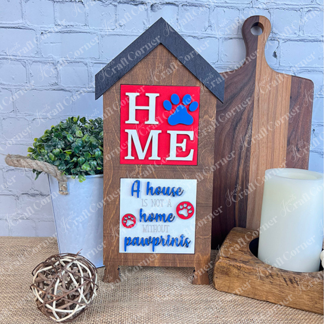 The "Dog Squares" sign by Janet's Craft Corner features wood cut-outs spelling "HOME" with a paw print for the "O" and the phrase, "A house is not a home without pawprints." Display it with plants, trays, candles, or decor.