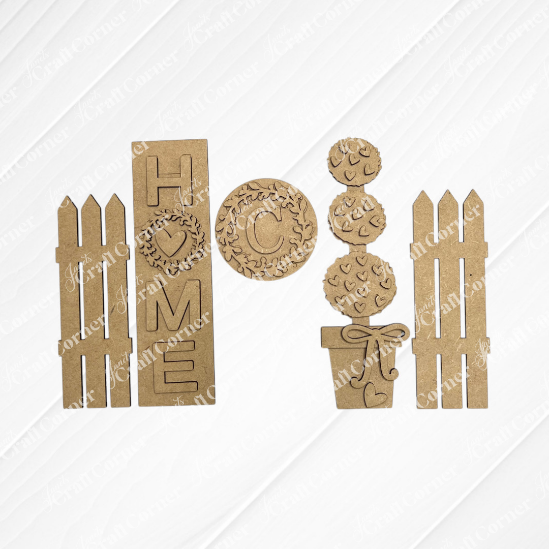 Janet's Craft Corner Home Interchangeable Porch Set includes two picket fences, a "HOME" sign, a wreath with "C," potted plant with hearts, and heart plaque—ideal for DIY seasonal home decor.