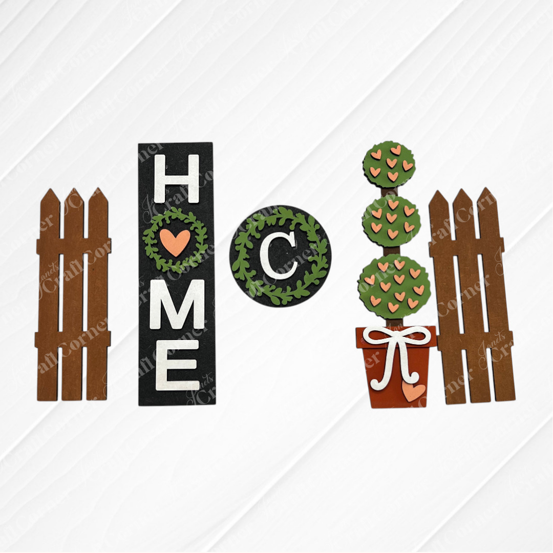 The Home Interchangeable Porch Set by Janet's Craft Corner includes wooden cutouts on a white background, with "HOME" inserts featuring a heart-shaped "O," a leafy round "C," brown fences, and a potted plant with heart-shaped leaves.