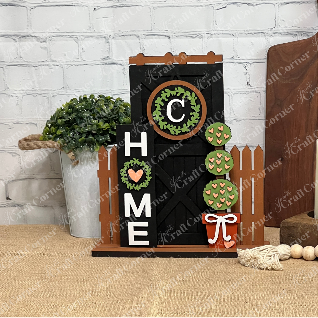 The Home Interchangeable Porch Set by Janet's Craft Corner features a wooden “HOME” sign with a wreath for the “O,” barn door, brown picket fence, greenery, and bow—perfect DIY seasonal decor for your porch or entryway.