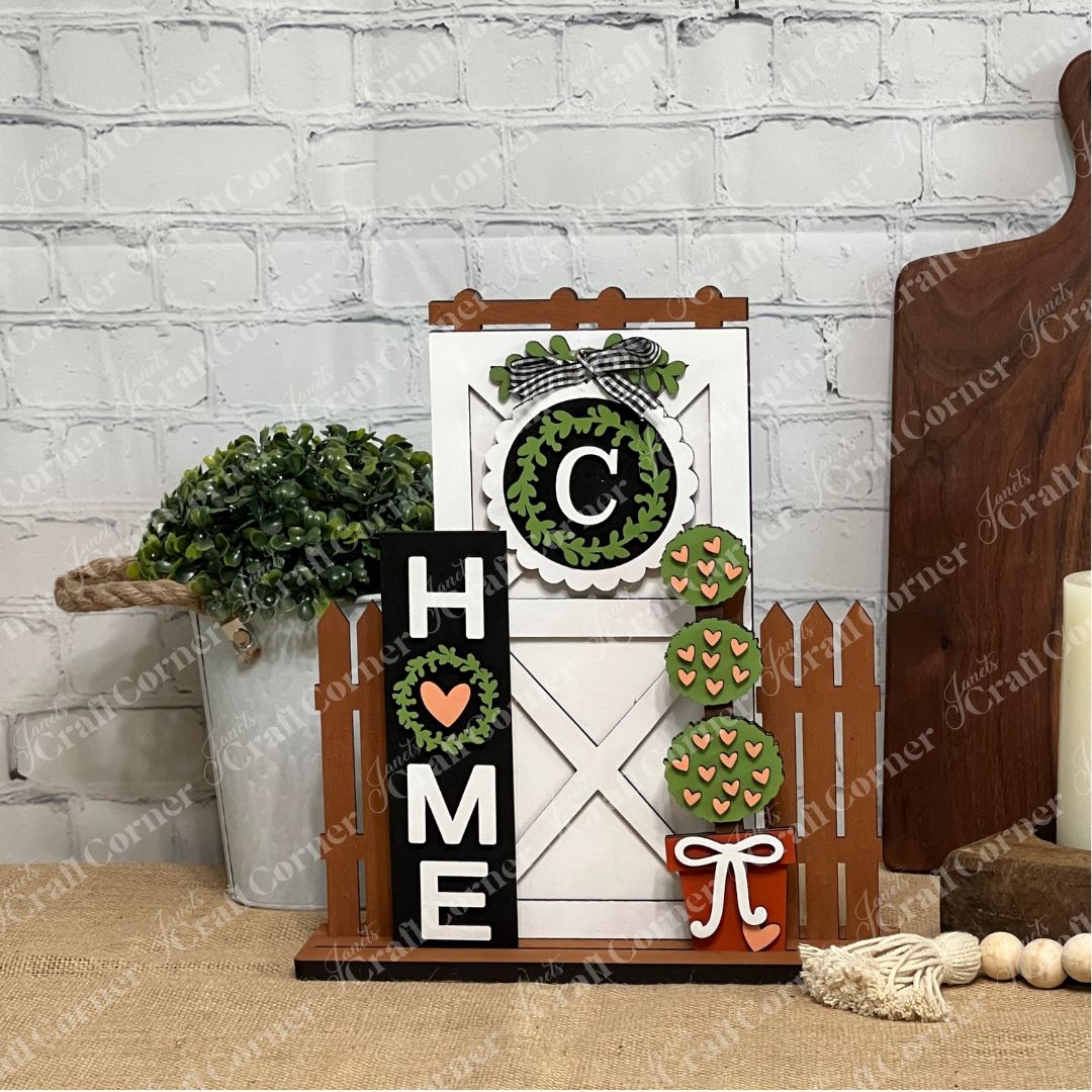 Janet's Craft Corner Home Interchangeable Porch Set features a farmhouse "HOME" sign with a heart for the "O," white barn door, picket fence, faux greenery, and mini trees—perfect DIY seasonal decor for your wooden surface.