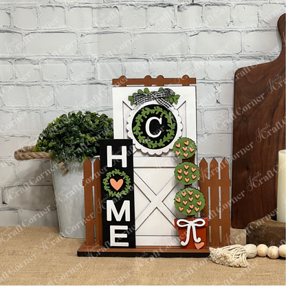 Janet's Craft Corner Home Interchangeable Porch Set features a farmhouse "HOME" sign with a heart for the "O," white barn door, picket fence, faux greenery, and mini trees—perfect DIY seasonal decor for your wooden surface.
